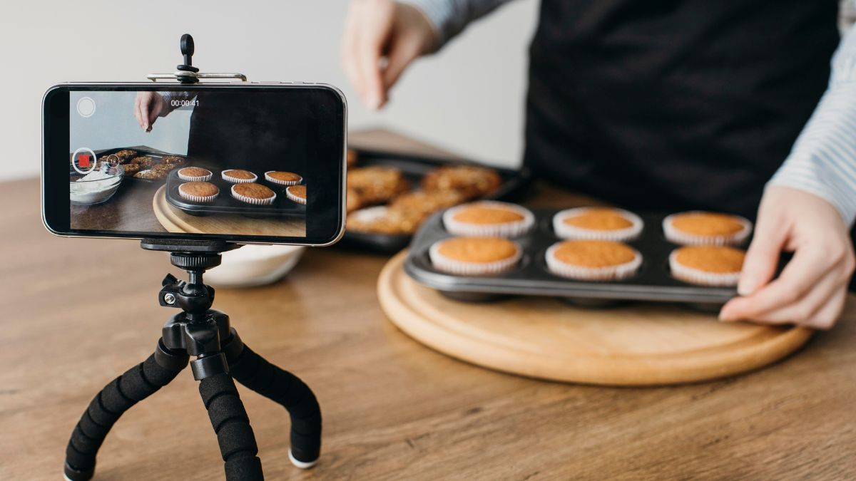 Person recording a video with a smartphone on a tripod, showing hands holding a tray of freshly baked cupcakes on a wooden board.