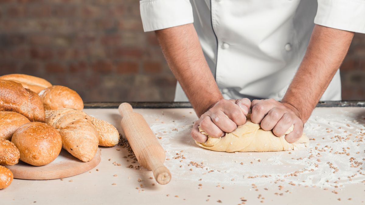 Person wearing a chef's uniform kneading dough on a floured counter, with a wooden rolling pin and assorted breads placed beside it.