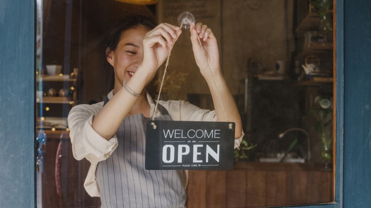 Smiling woman in an apron hangs a "Welcome, we are open" sign on the glass door of a small business, seen from outside, suggesting a friendly and inviting atmosphere.