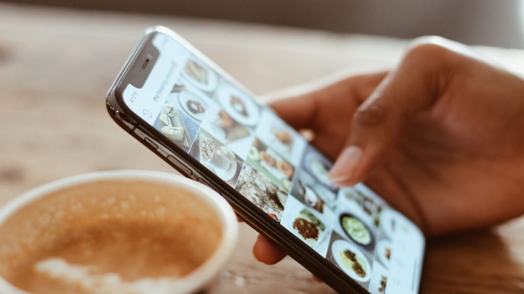 Hand holding a smartphone displaying photos of food dishes, with a cup of coffee with foam in the foreground on a wooden table.