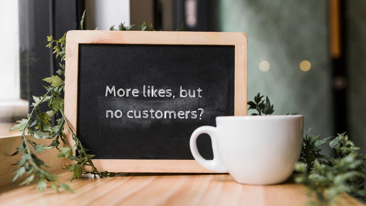 Blackboard with a wooden frame on a wooden table, with the phrase "More likes, but no customers?" written in white; next to it, a white cup and decorative leafy branches.