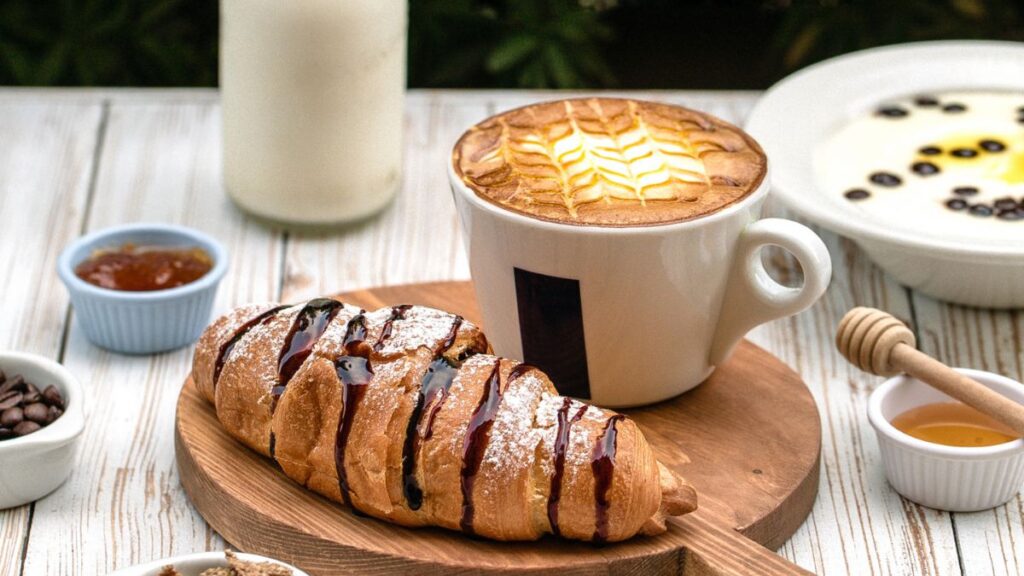 A freshly baked chocolate-drizzled croissant on a wooden board, served with a cup of cappuccino topped with elegant latte art. Surrounding the main dish are small bowls of jam, honey, coffee beans, and yogurt, creating a cozy and inviting breakfast setup on a rustic white wooden table.