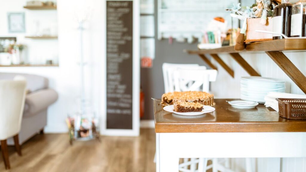 A slice of chocolate and caramel cake on a white plate sits on a wooden counter in a cozy, well-lit café with white furniture and a blurred blackboard menu in the background.