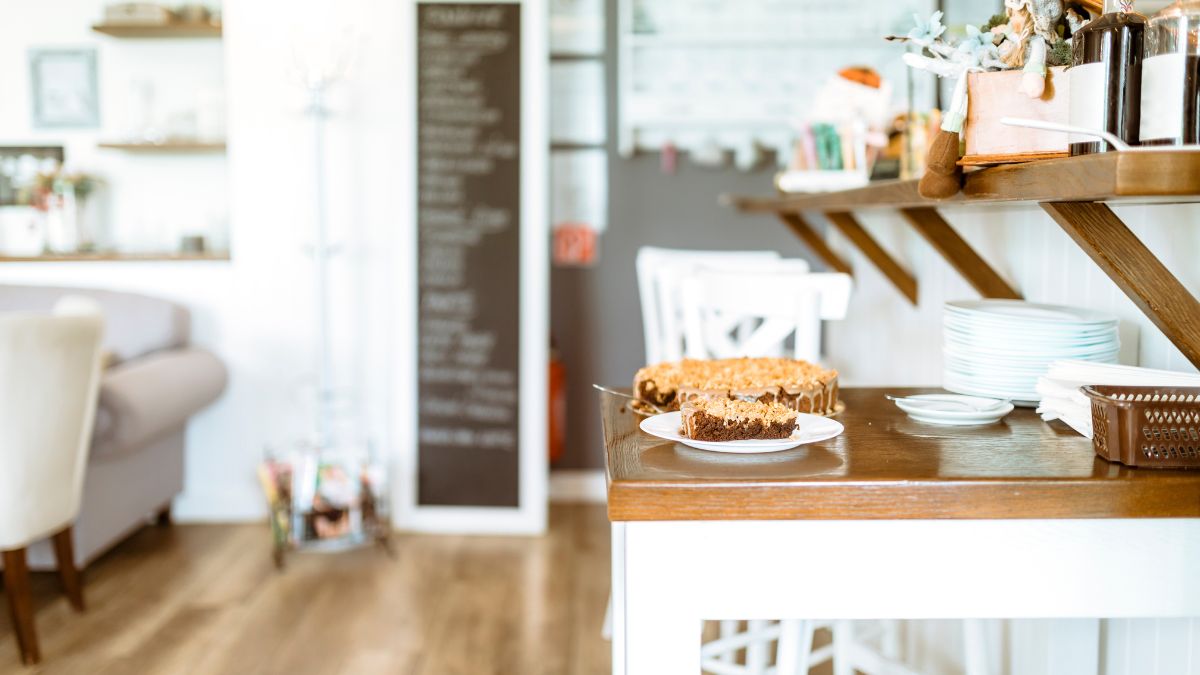 A slice of chocolate and caramel cake on a white plate sits on a wooden counter in a cozy, well-lit café with white furniture and a blurred blackboard menu in the background.